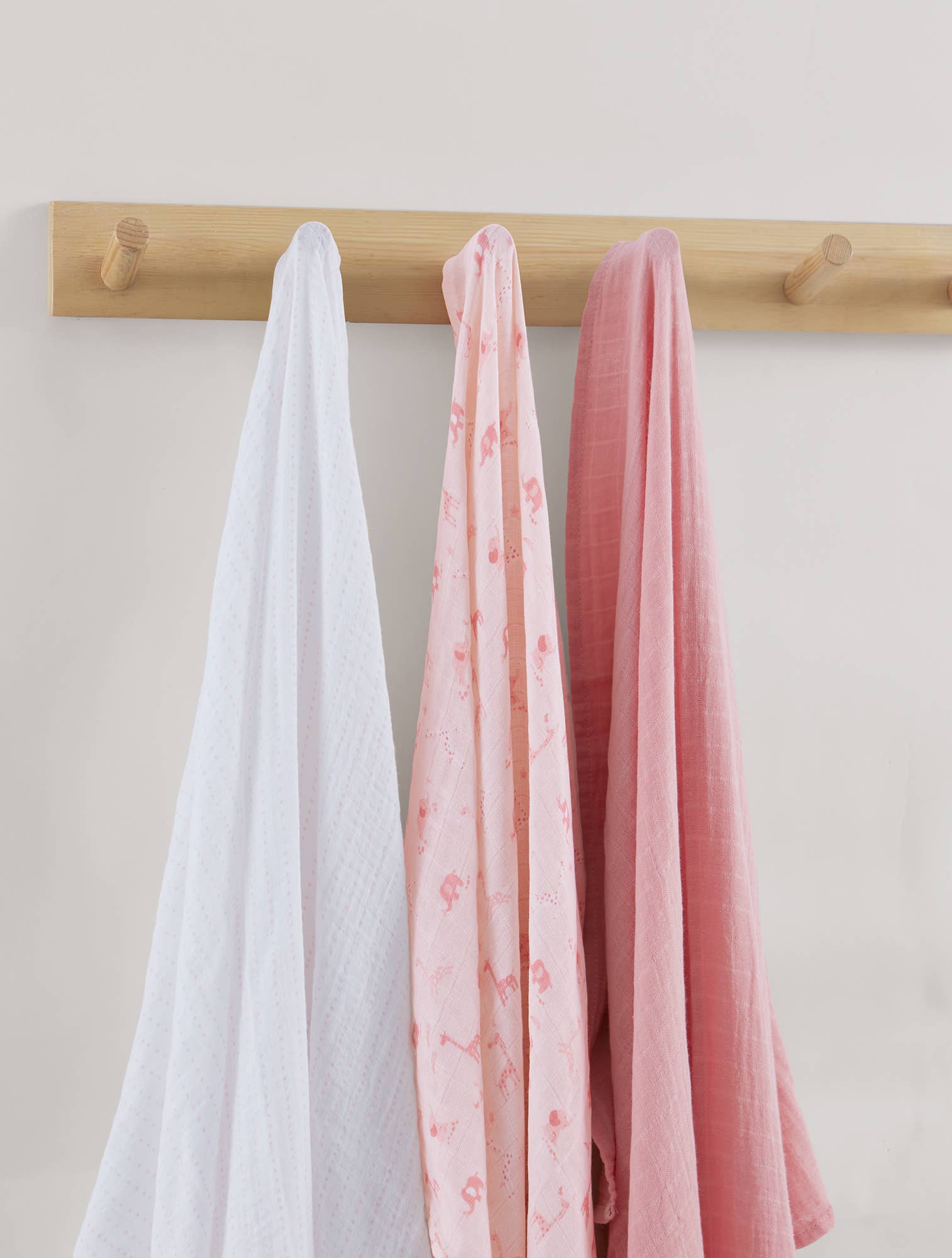 Three swaddles in white, pink, and red hanging on a wooden rod against a light background.