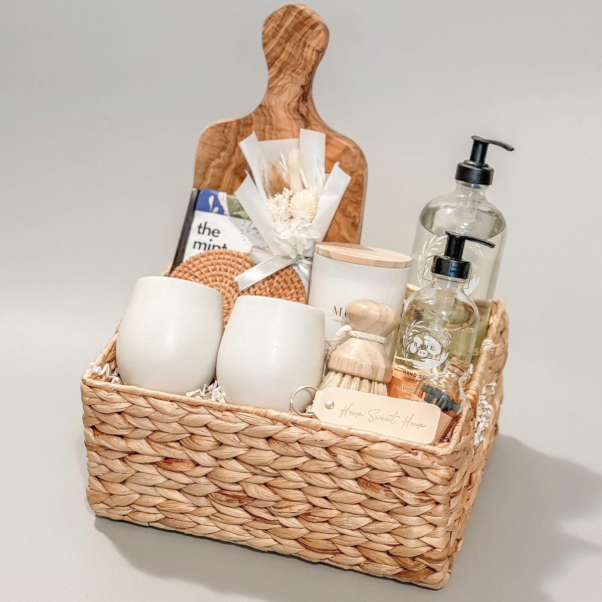 Luxury housewarming gift basket featuring ceramic mugs, natural soap, hand lotion, a wooden dish brush, candle, dried florals, and a rustic charcuterie board in a woven basket
