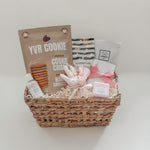 Rosalie self-care gift basket by Muse Baskets and Co. shown straight on, featuring YVR Cookie chocolate chip crisps, Handfuel everything bagel cashews, a So Luxury blush soak, a Fresh Wife black raspberry bath bomb and hand lotion, and a white and pink preserved floral bouquet in a woven wicker basket, a soft self-care gift for her