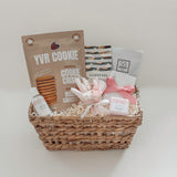 Rosalie self-care gift basket by Muse Baskets and Co. shown straight on, featuring YVR Cookie chocolate chip crisps, Handfuel everything bagel cashews, a So Luxury blush soak, a Fresh Wife black raspberry bath bomb and hand lotion, and a white and pink preserved floral bouquet in a woven wicker basket, a soft self-care gift for her