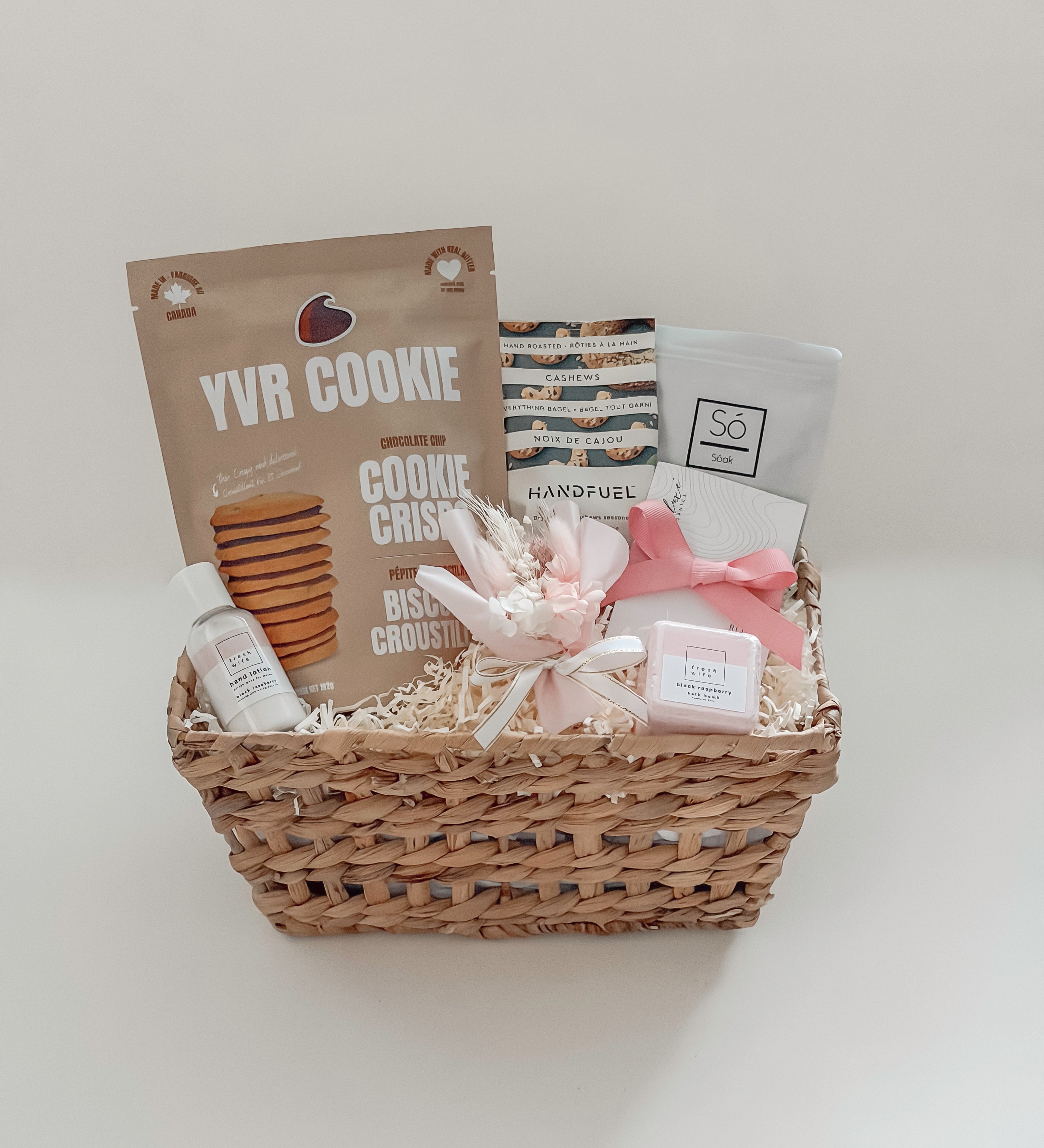 Rosalie self-care gift basket by Muse Baskets and Co. shown straight on, featuring YVR Cookie chocolate chip crisps, Handfuel everything bagel cashews, a So Luxury blush soak, a Fresh Wife black raspberry bath bomb and hand lotion, and a white and pink preserved floral bouquet in a woven wicker basket, a soft self-care gift for her