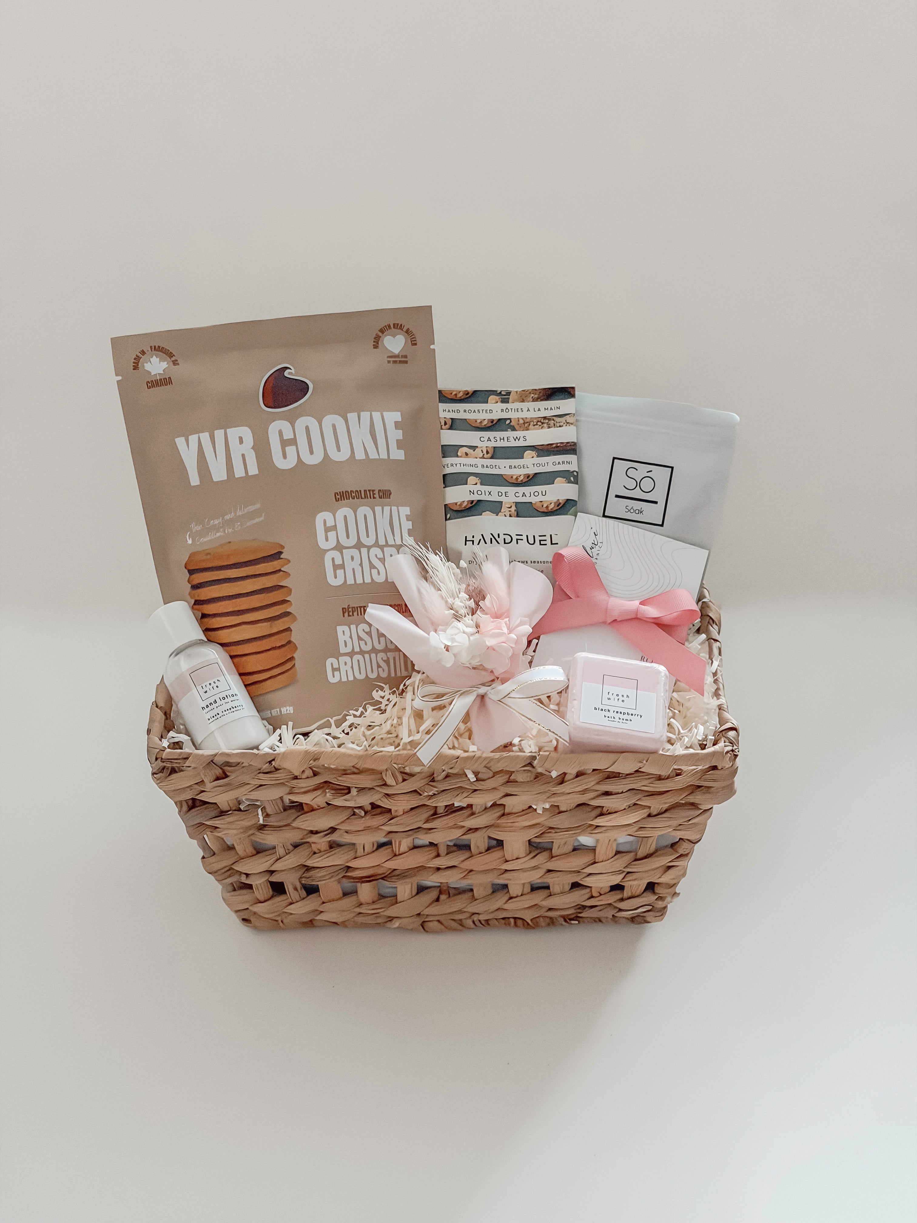 Rosalie self-care gift basket by Muse Baskets and Co. shown straight on, featuring YVR Cookie chocolate chip crisps, Handfuel everything bagel cashews, a So Luxury blush soak, a Fresh Wife black raspberry bath bomb and hand lotion, and a white and pink preserved floral bouquet in a woven wicker basket, a soft self-care gift for her