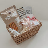 Rosalie gift basket by Muse Baskets and Co. photographed at an angle, with YVR Cookie crisps, Handfuel cashews, a So Luxury blush soak, a Sealuxe rose face mask tied with pink ribbon, a Fresh Wife black raspberry bath bomb and hand lotion, and preserved white and pink florals in a wicker basket from Canadian and women-owned makers