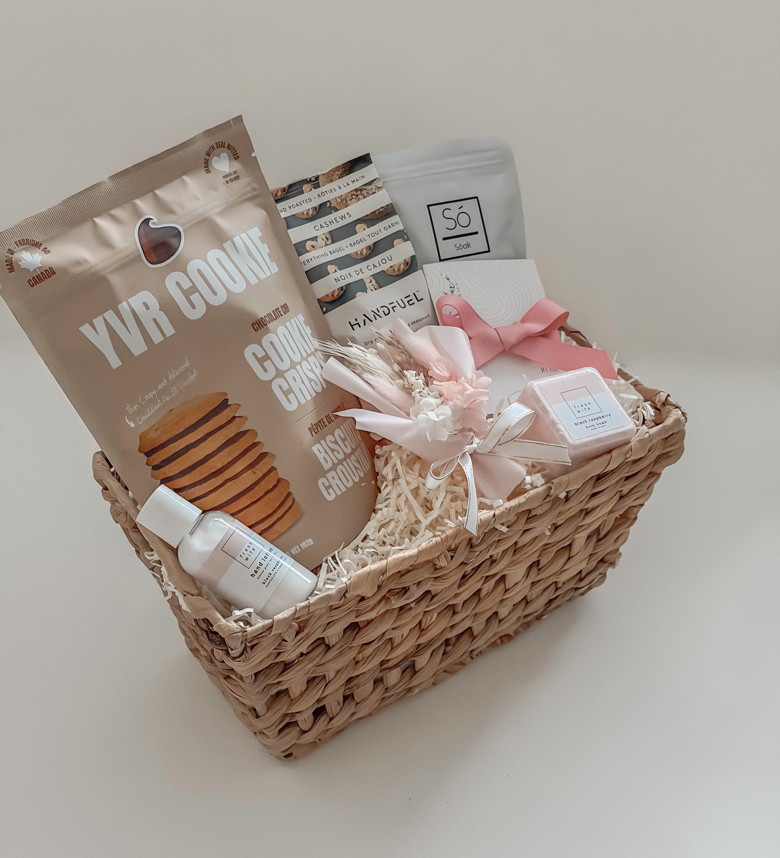 Rosalie gift basket by Muse Baskets and Co. photographed at an angle, with YVR Cookie crisps, Handfuel cashews, a So Luxury blush soak, a Sealuxe rose face mask tied with pink ribbon, a Fresh Wife black raspberry bath bomb and hand lotion, and preserved white and pink florals in a wicker basket from Canadian and women-owned makers