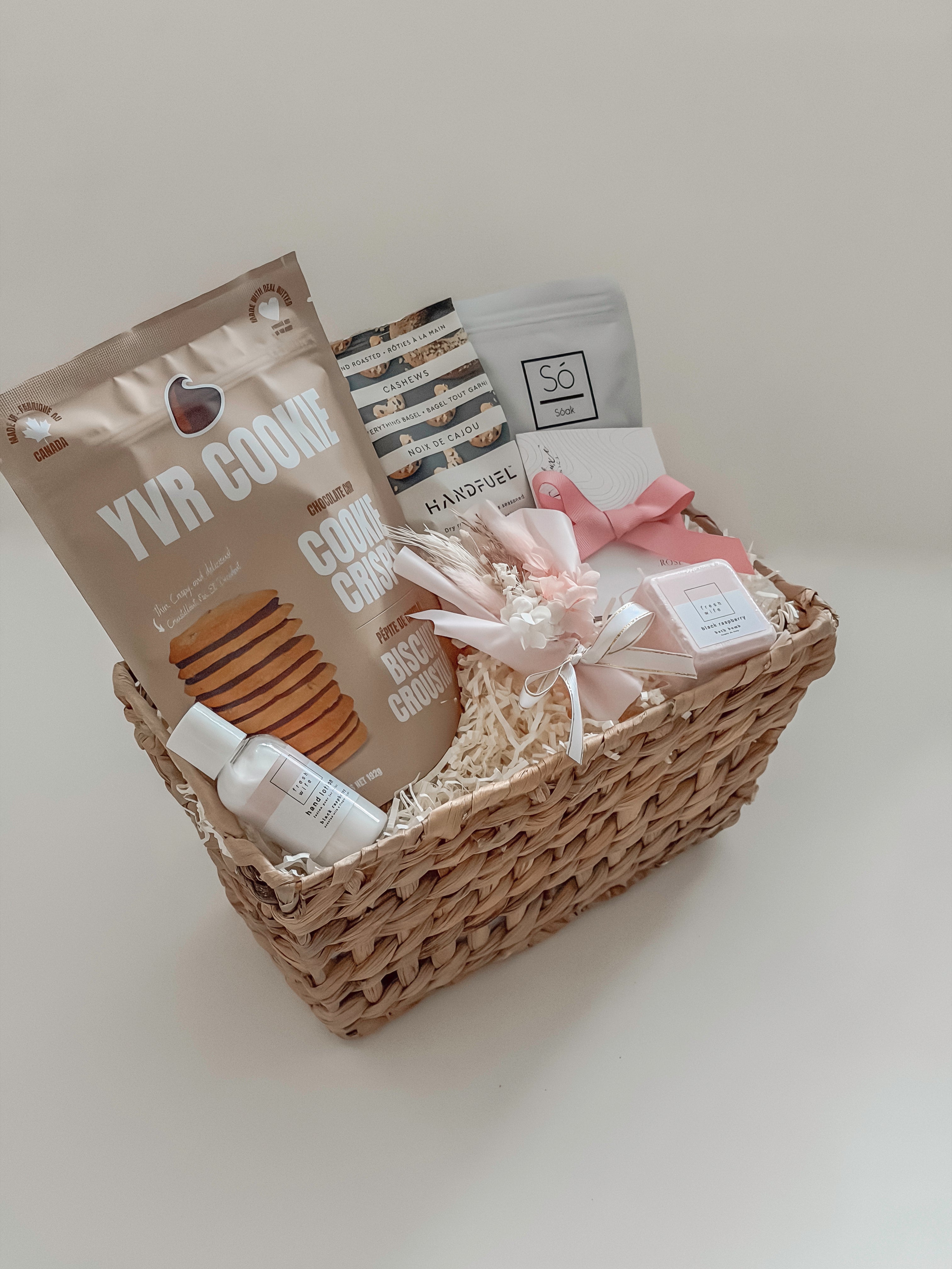 Rosalie gift basket by Muse Baskets and Co. photographed at an angle, with YVR Cookie crisps, Handfuel cashews, a So Luxury blush soak, a Sealuxe rose face mask tied with pink ribbon, a Fresh Wife black raspberry bath bomb and hand lotion, and preserved white and pink florals in a wicker basket from Canadian and women-owned makers
