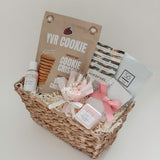 Rosalie luxury gift basket for her by Muse Baskets and Co. viewed from above, showing YVR Cookie chocolate chip crisps, Handfuel everything bagel cashews, a So Luxury blush soak, a Sealuxe rose face mask, a Fresh Wife black raspberry bath bomb and hand lotion, and a preserved floral bouquet in shades of white and pink in a woven wicker basket