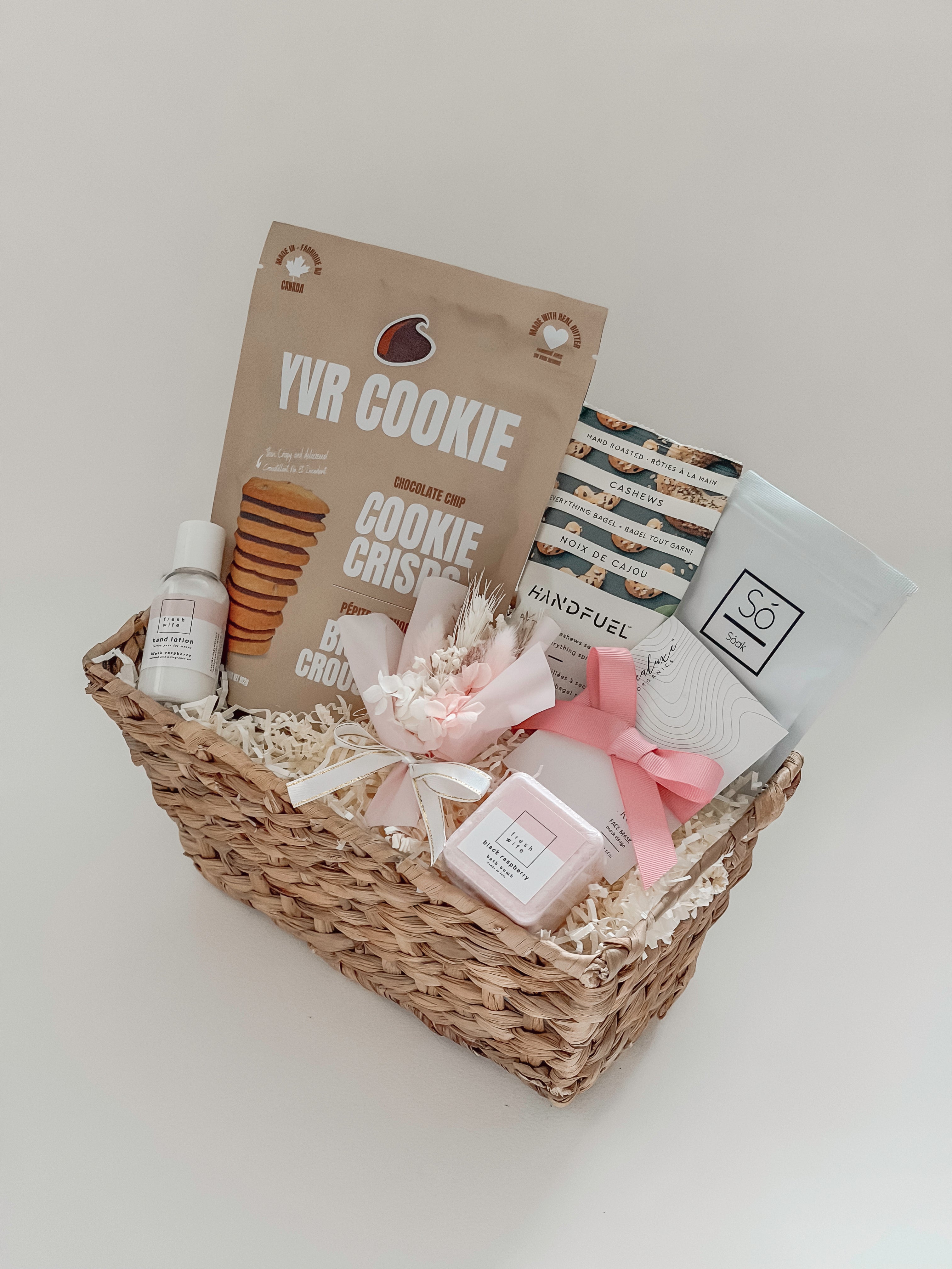 Rosalie luxury gift basket for her by Muse Baskets and Co. viewed from above, showing YVR Cookie chocolate chip crisps, Handfuel everything bagel cashews, a So Luxury blush soak, a Sealuxe rose face mask, a Fresh Wife black raspberry bath bomb and hand lotion, and a preserved floral bouquet in shades of white and pink in a woven wicker basket