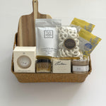 Thanksgiving gift basket by Muse Baskets and Co. with acacia wood board, raw honey, tea, candle, white chocolate pretzels and bath items in a woven basket.