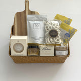 Thanksgiving gift basket by Muse Baskets and Co. with acacia wood board, raw honey, tea, candle, white chocolate pretzels and bath items in a woven basket.
