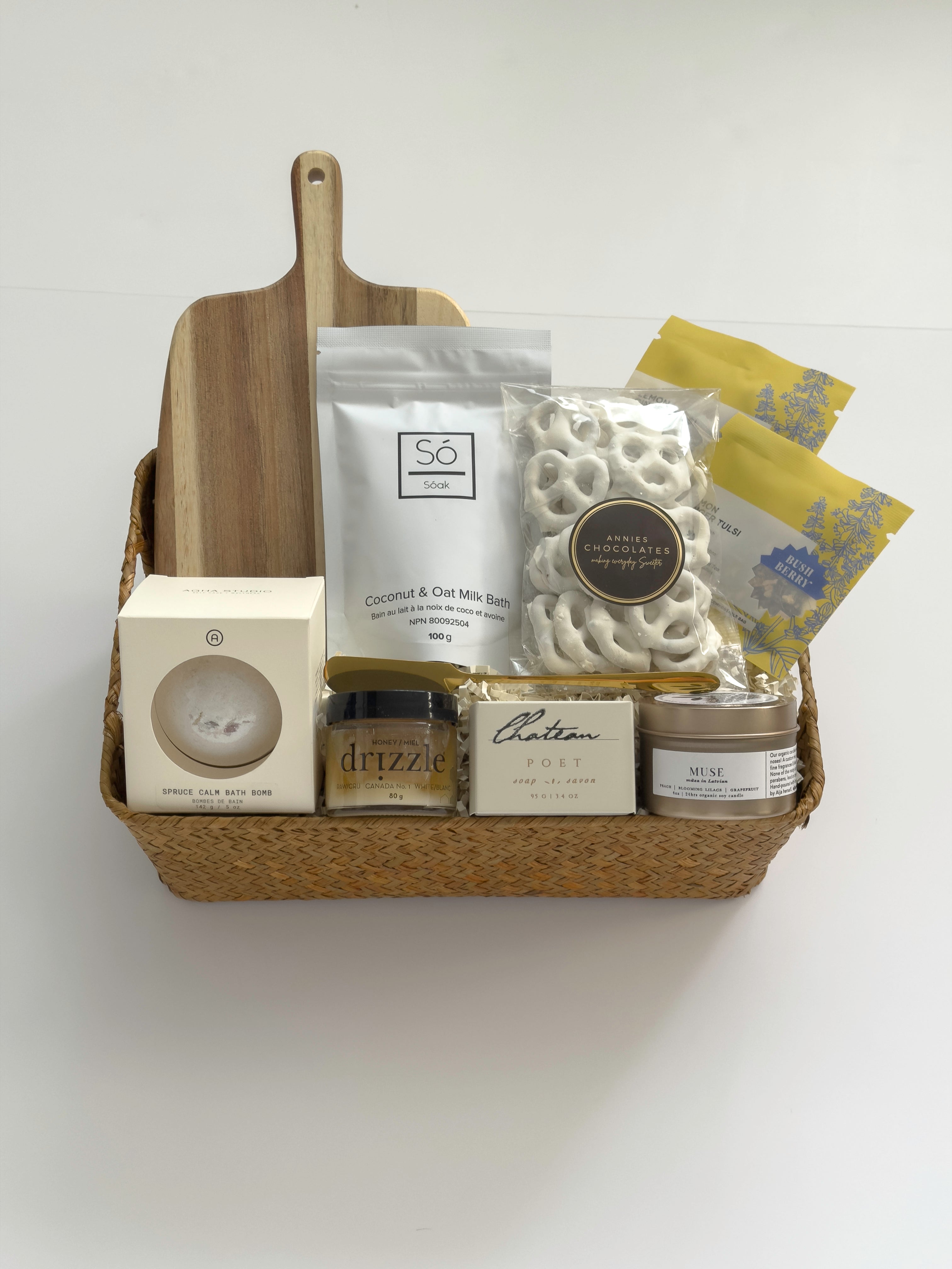 Thanksgiving gift basket by Muse Baskets and Co. with acacia wood board, raw honey, tea, candle, white chocolate pretzels and bath items in a woven basket.
