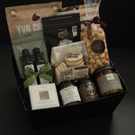 The Gathered Home gift basket by Muse Baskets and Co. shown from above, featuring YVR Cookie crisps, Roger's chocolate covered espresso beans, Annie's chocolate popcorn, Bliss Pasticinni cookies, infused olive oil and balsamic, Pear Riesling wine jam, SaltSpring candied jalapeño relish, a porcelain dipping dish, and a Noel & Co. candle in a black gift box, a refined housewarming gift, realtor closing gift, and possession day gift.