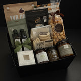 The Gathered Home gift basket by Muse Baskets and Co. shown from above, featuring YVR Cookie crisps, Roger's chocolate covered espresso beans, Annie's chocolate popcorn, Bliss Pasticinni cookies, infused olive oil and balsamic, Pear Riesling wine jam, SaltSpring candied jalapeño relish, a porcelain dipping dish, and a Noel & Co. candle in a black gift box, a refined housewarming gift, realtor closing gift, and possession day gift.