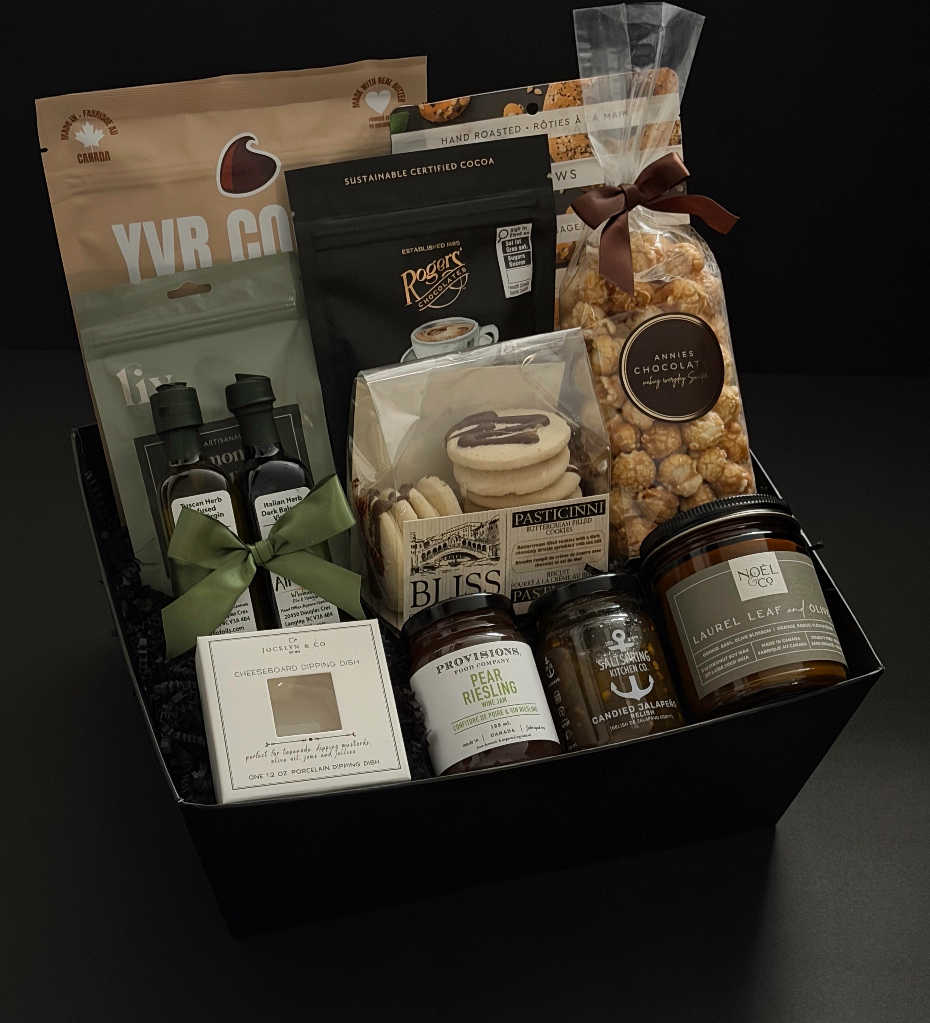 The Gathered Home gift basket by Muse Baskets and Co. shown from above, featuring YVR Cookie crisps, Roger's chocolate covered espresso beans, Annie's chocolate popcorn, Bliss Pasticinni cookies, infused olive oil and balsamic, Pear Riesling wine jam, SaltSpring candied jalapeño relish, a porcelain dipping dish, and a Noel & Co. candle in a black gift box, a refined housewarming gift, realtor closing gift, and possession day gift.