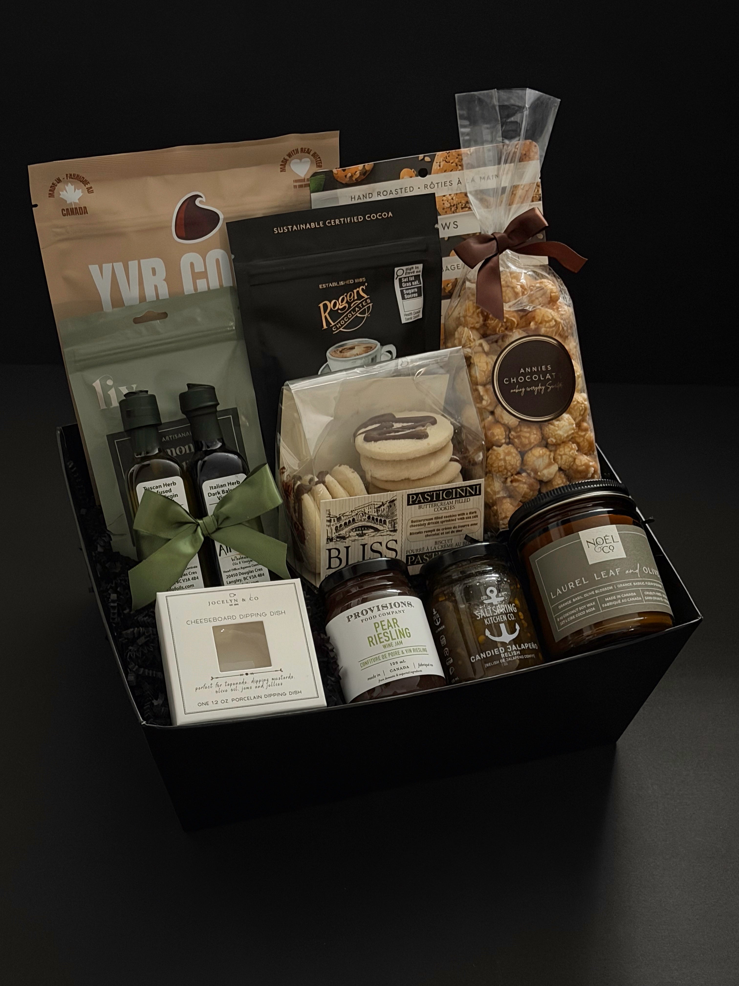 The Gathered Home gift basket by Muse Baskets and Co. shown from above, featuring YVR Cookie crisps, Roger's chocolate covered espresso beans, Annie's chocolate popcorn, Bliss Pasticinni cookies, infused olive oil and balsamic, Pear Riesling wine jam, SaltSpring candied jalapeño relish, a porcelain dipping dish, and a Noel & Co. candle in a black gift box, a refined housewarming gift, realtor closing gift, and possession day gift.