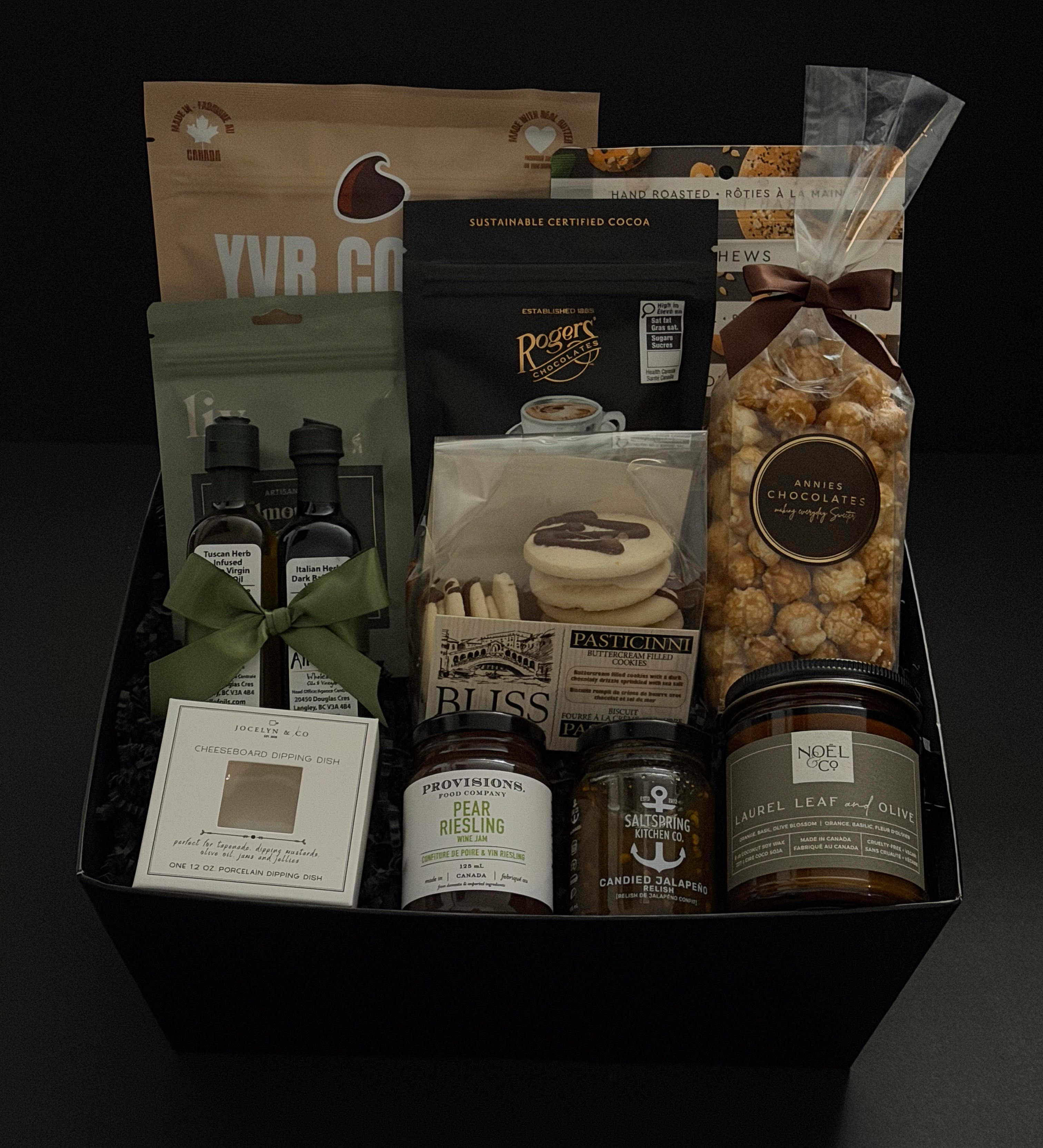 The Gathered Home gift basket by Muse Baskets and Co. photographed straight on, with YVR Cookie crisps, Roger's espresso beans, Annie's chocolate popcorn, Bliss buttercream cookies, All of Oils olive oil and balsamic, Provisions wine jam, SaltSpring candied jalapeño relish, a Jocelyn & Co. dipping dish, and a Laurel Leaf and Olive candle by Noel & Co. in a black box, a curated client and corporate housewarming gift from Canadian makers.