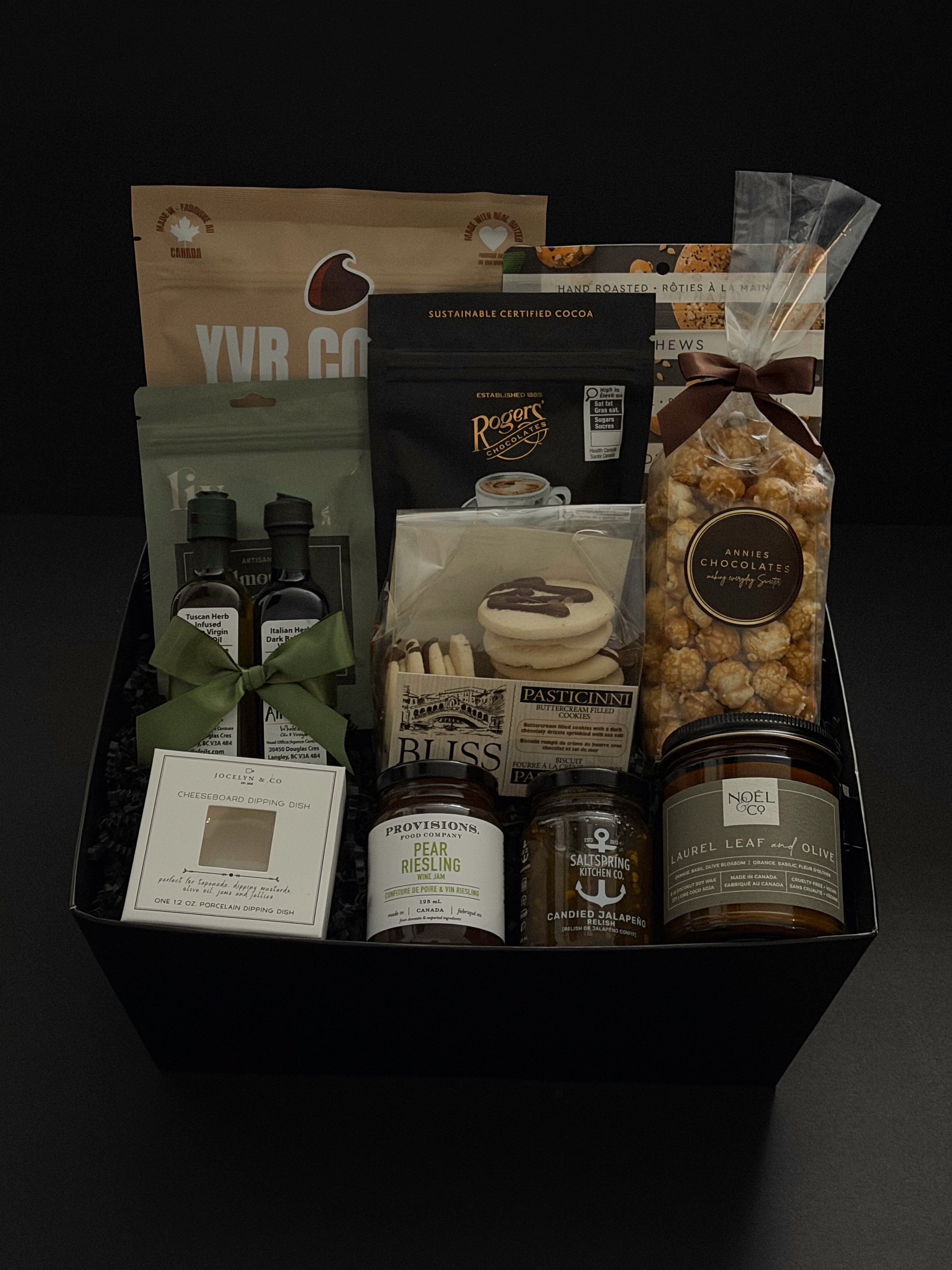 The Gathered Home gift basket by Muse Baskets and Co. photographed straight on, with YVR Cookie crisps, Roger's espresso beans, Annie's chocolate popcorn, Bliss buttercream cookies, All of Oils olive oil and balsamic, Provisions wine jam, SaltSpring candied jalapeño relish, a Jocelyn & Co. dipping dish, and a Laurel Leaf and Olive candle by Noel & Co. in a black box, a curated client and corporate housewarming gift from Canadian makers.