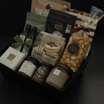 The Gathered Home gift basket by Muse Baskets and Co. viewed close up at an angle, showing YVR Cookie crisps, Roger's chocolate covered espresso beans, Annie's chocolate popcorn, Bliss Pasticinni cookies, infused olive oil and balsamic, Pear Riesling wine jam, SaltSpring candied jalapeño relish, a porcelain dipping dish, and a Noel & Co. candle in a black gift box, a refined realtor closing gift, possession day gift, and client gift across Canada.