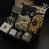The Gathered Home gift basket by Muse Baskets and Co. viewed close up at an angle, showing YVR Cookie crisps, Roger's chocolate covered espresso beans, Annie's chocolate popcorn, Bliss Pasticinni cookies, infused olive oil and balsamic, Pear Riesling wine jam, SaltSpring candied jalapeño relish, a porcelain dipping dish, and a Noel & Co. candle in a black gift box, a refined realtor closing gift, possession day gift, and client gift across Canada.