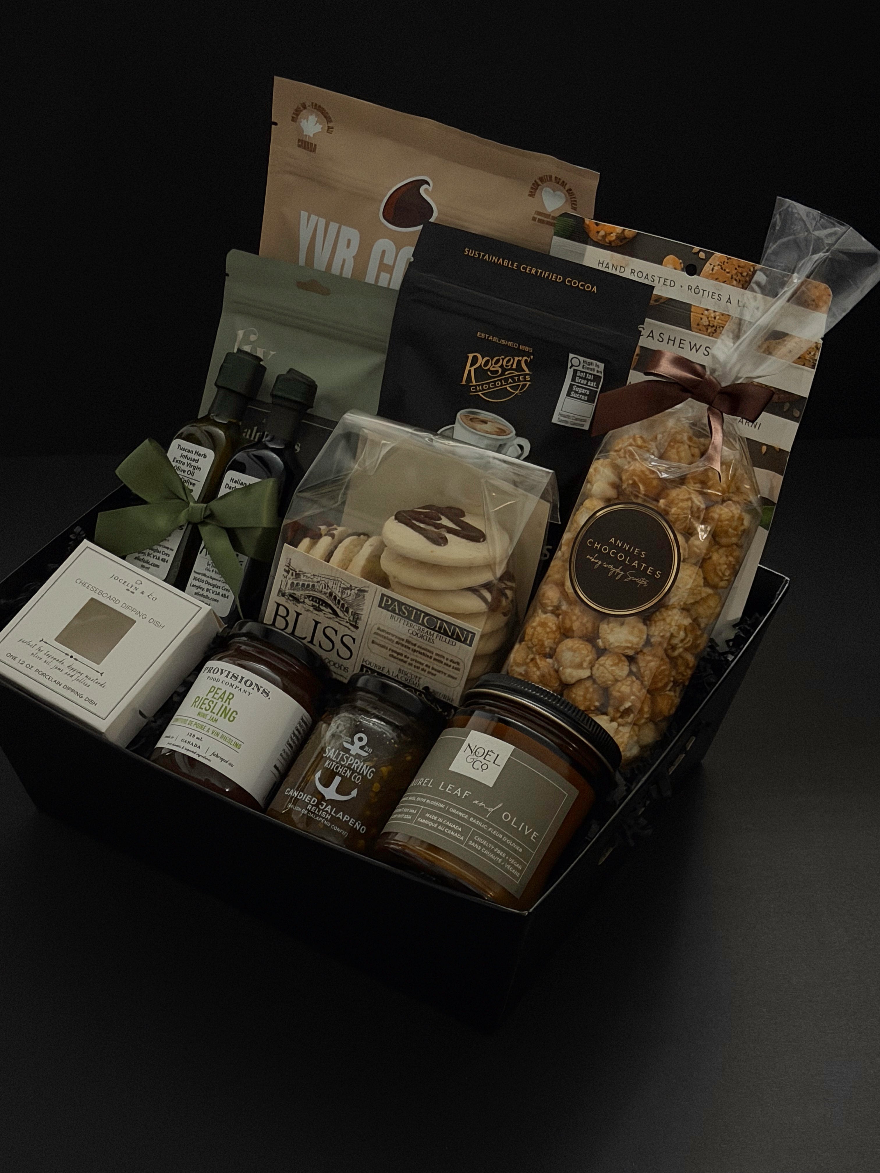 The Gathered Home gift basket by Muse Baskets and Co. viewed close up at an angle, showing YVR Cookie crisps, Roger's chocolate covered espresso beans, Annie's chocolate popcorn, Bliss Pasticinni cookies, infused olive oil and balsamic, Pear Riesling wine jam, SaltSpring candied jalapeño relish, a porcelain dipping dish, and a Noel & Co. candle in a black gift box, a refined realtor closing gift, possession day gift, and client gift across Canada.