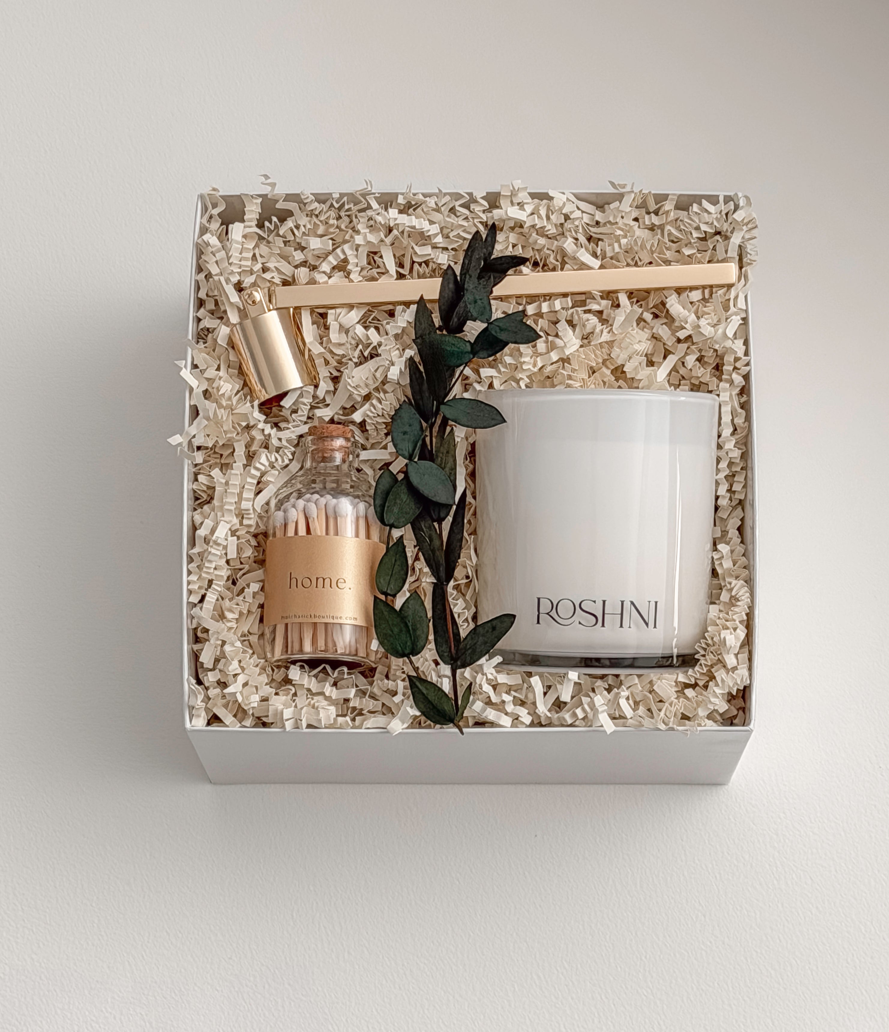 Lumière gift box by Muse Baskets and Co. shown from above, featuring a white Roshni candle, a gold candle snuffer, white tipped home matches in a glass jar, and a preserved eucalyptus stem arranged on natural fill in a white box, a refined realtor closing gift, possession day gift, and corporate gift.