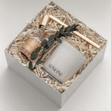 Lumière gift box by Muse Baskets and Co. viewed from the side, showing a white Roshni candle, gold candle snuffer, glass jar of white tipped home matches, and preserved eucalyptus on natural fill in a white box, a curated possession day gift, realtor closing gift, and corporate gift with Canada-wide delivery.
