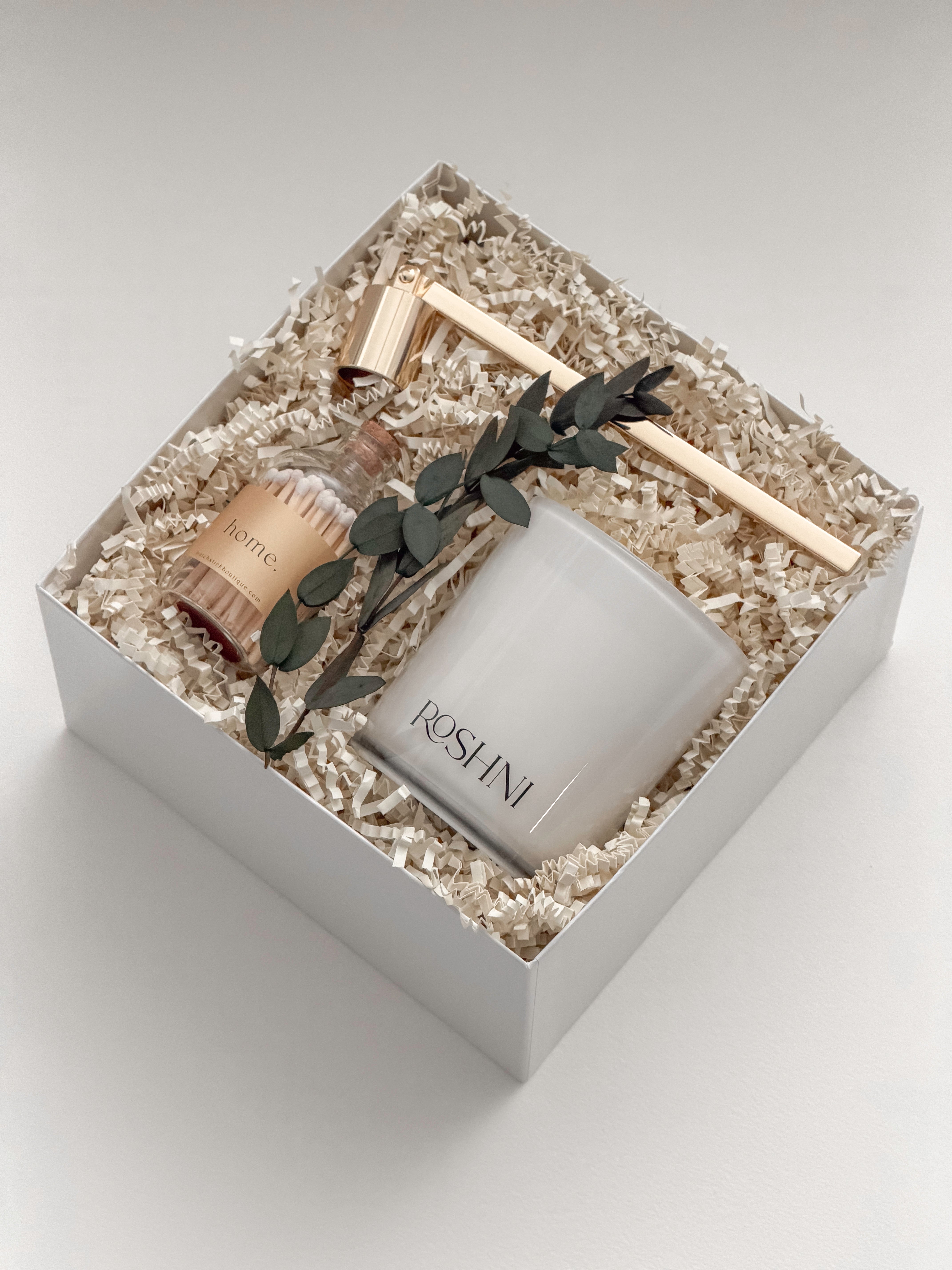 Lumière gift box by Muse Baskets and Co. viewed from the side, showing a white Roshni candle, gold candle snuffer, glass jar of white tipped home matches, and preserved eucalyptus on natural fill in a white box, a curated possession day gift, realtor closing gift, and corporate gift with Canada-wide delivery.