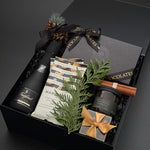 A luxury gift hamper box set on a dark grey background, featuring a bottle of Freixenet sparkling wine, handcrafted dessert chocolates, hand-roasted cashews, luxurious candle and a small wrapped gift box with chocolate almonds.