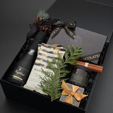 A luxury gift hamper box set on a dark grey background, featuring a bottle of Freixenet sparkling wine, handcrafted dessert chocolates, hand-roasted cashews, luxurious candle and a small wrapped gift box with chocolate almonds.