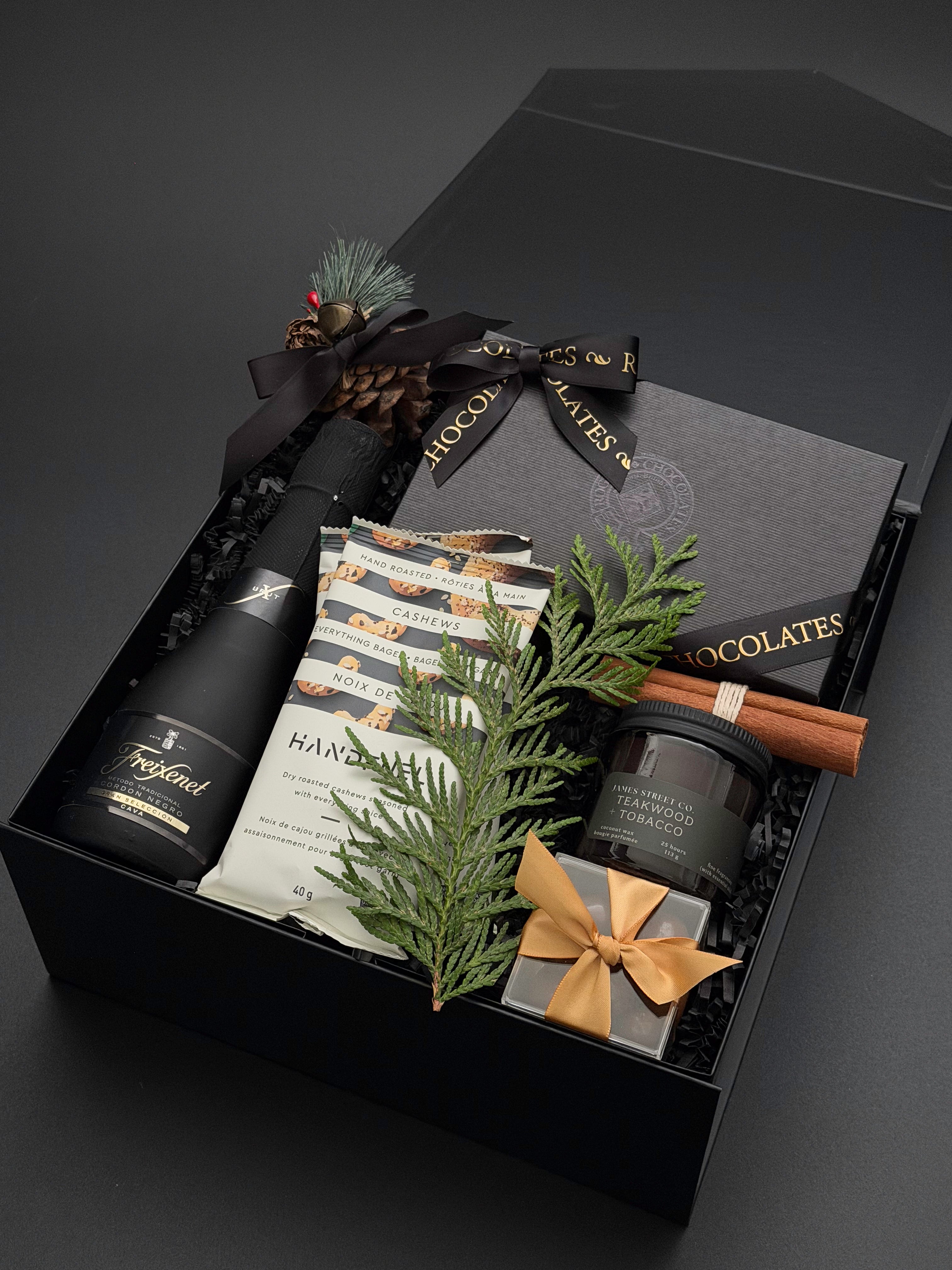 A luxury gift hamper box set on a dark grey background, featuring a bottle of Freixenet sparkling wine, handcrafted dessert chocolates, hand-roasted cashews, luxurious candle and a small wrapped gift box with chocolate almonds.