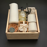 A curated gift set in a light natural wood tray, featuring a tall matte white ceramic vessel, a bottle of hand soap, two small white ceramic cups, a wrapped soap bar tied with a cream ribbon, a round brown scrub, and a small green tag, all arranged on a dark gray background.