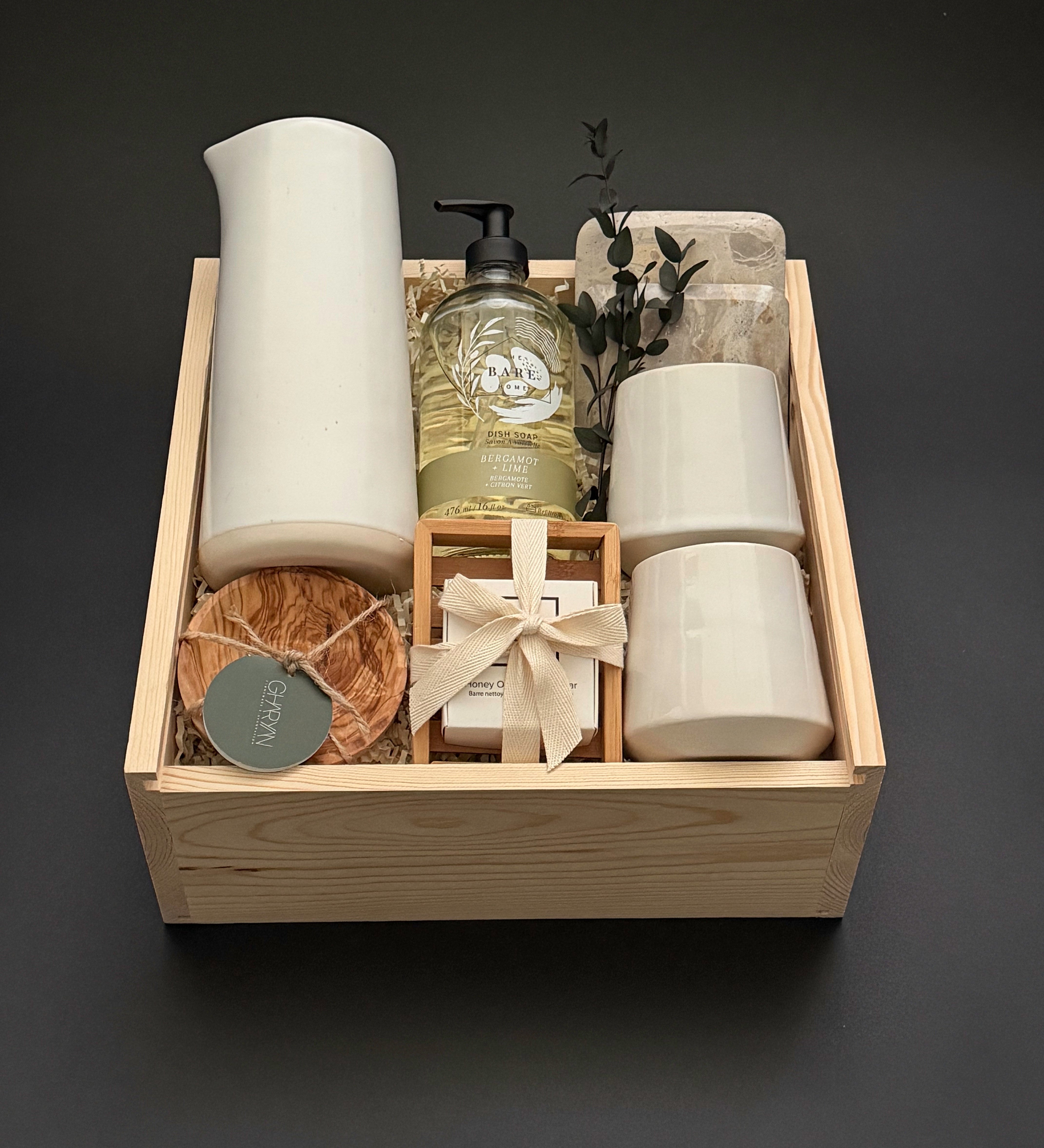 A curated gift set in a light natural wood tray, featuring a tall matte white ceramic vessel, a bottle of hand soap, two small white ceramic cups, a wrapped soap bar tied with a cream ribbon, a round brown scrub, and a small green tag, all arranged on a dark gray background.