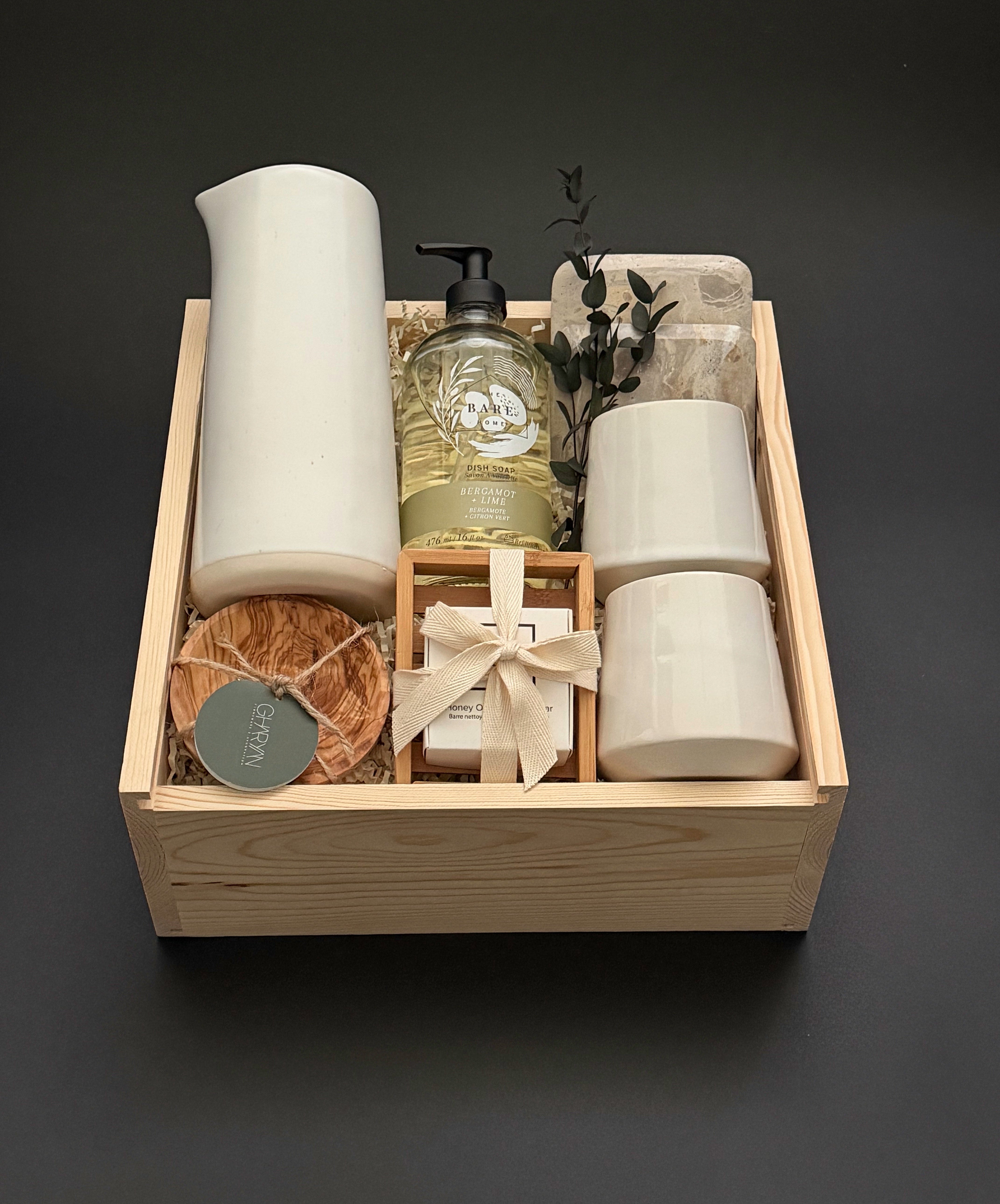 A curated gift set in a light natural wood tray, featuring a tall matte white ceramic vessel, a bottle of hand soap, two small white ceramic cups, a wrapped soap bar tied with a cream ribbon, a round brown scrub, and a small green tag, all arranged on a dark gray background.