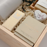 A three-quarter view of The West Village gift box from Muse Baskets and Co., a Vancouver gifting studio, containing a white Fable ceramic carafe, an Agha Studio Nordic Spa face and body savon, a heart-shaped Reverie bath bomb, a Vancouver Candle Co. Silentium candle, and a Laura Slack chocolate bar, finished with eucalyptus. A possession day gift and homeowner welcome gift.