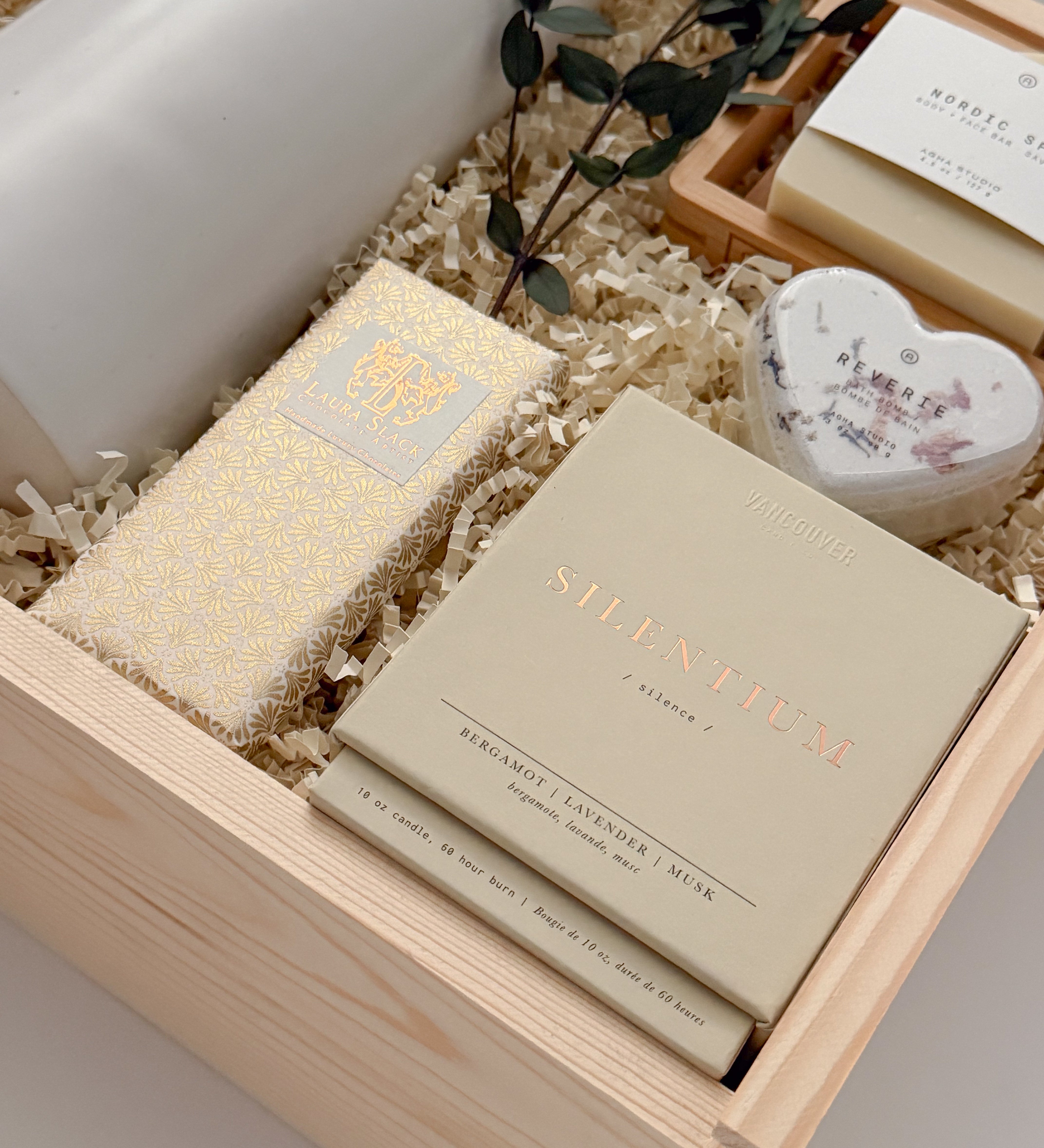 A three-quarter view of The West Village gift box from Muse Baskets and Co., a Vancouver gifting studio, containing a white Fable ceramic carafe, an Agha Studio Nordic Spa face and body savon, a heart-shaped Reverie bath bomb, a Vancouver Candle Co. Silentium candle, and a Laura Slack chocolate bar, finished with eucalyptus. A possession day gift and homeowner welcome gift.
