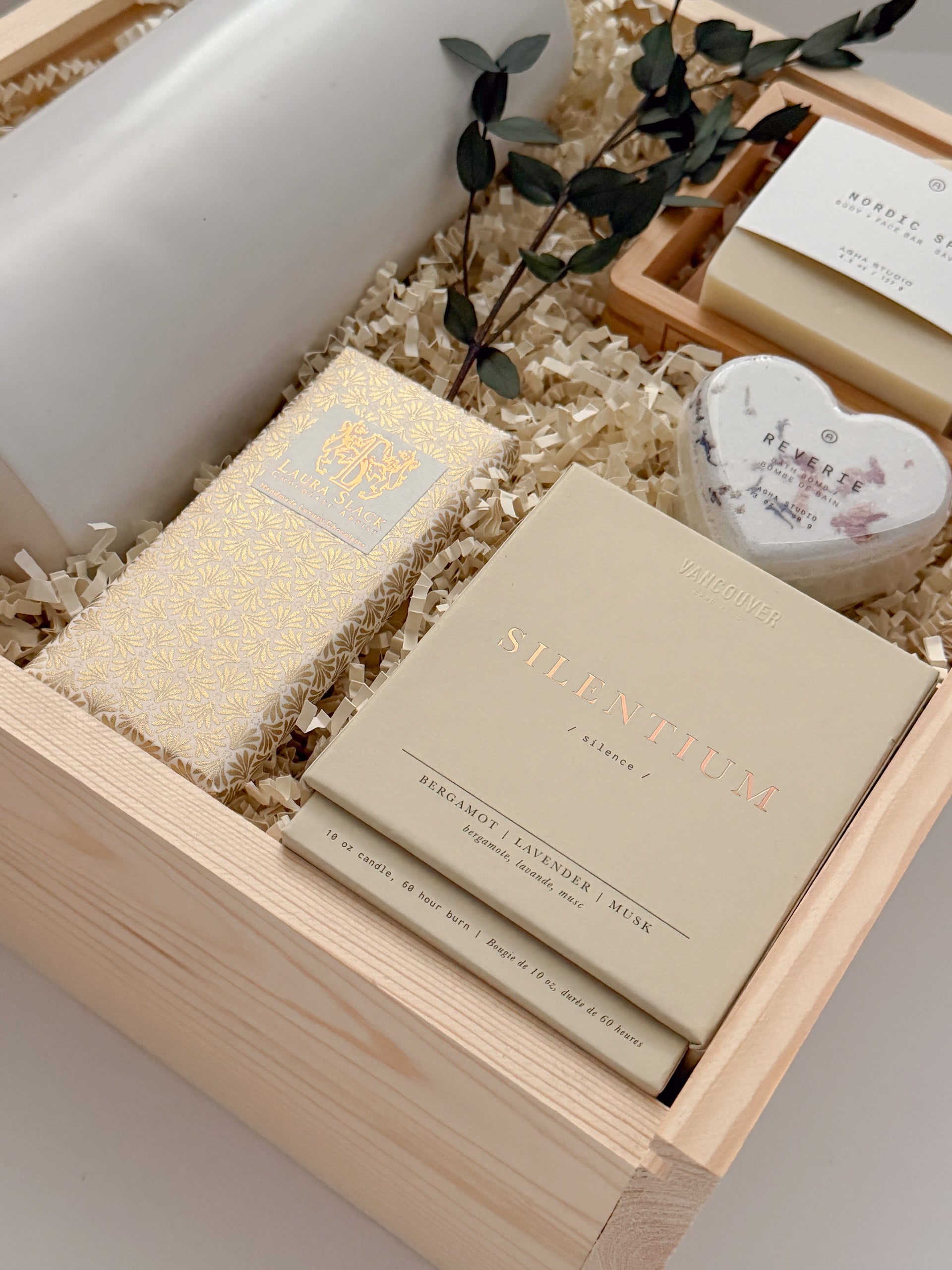 A three-quarter view of The West Village gift box from Muse Baskets and Co., a Vancouver gifting studio, containing a white Fable ceramic carafe, an Agha Studio Nordic Spa face and body savon, a heart-shaped Reverie bath bomb, a Vancouver Candle Co. Silentium candle, and a Laura Slack chocolate bar, finished with eucalyptus. A possession day gift and homeowner welcome gift.