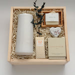 Overhead flat lay of The West Village by Muse Baskets and Co., showing a speckled white Fable carafe standing upright beside a Laura Slack artisan chocolate bar, a Vancouver Candle Co. Silentium candle, a Reverie heart bath bomb, and an Agha Studio Nordic Spa savon in a So Luxury bamboo dish, arranged in a Signature Luxe wood gift box. A luxury realtor closing gift and corporate gift.
