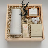 Overhead flat lay of The West Village by Muse Baskets and Co., showing a speckled white Fable carafe standing upright beside a Laura Slack artisan chocolate bar, a Vancouver Candle Co. Silentium candle, a Reverie heart bath bomb, and an Agha Studio Nordic Spa savon in a So Luxury bamboo dish, arranged in a Signature Luxe wood gift box. A luxury realtor closing gift and corporate gift.