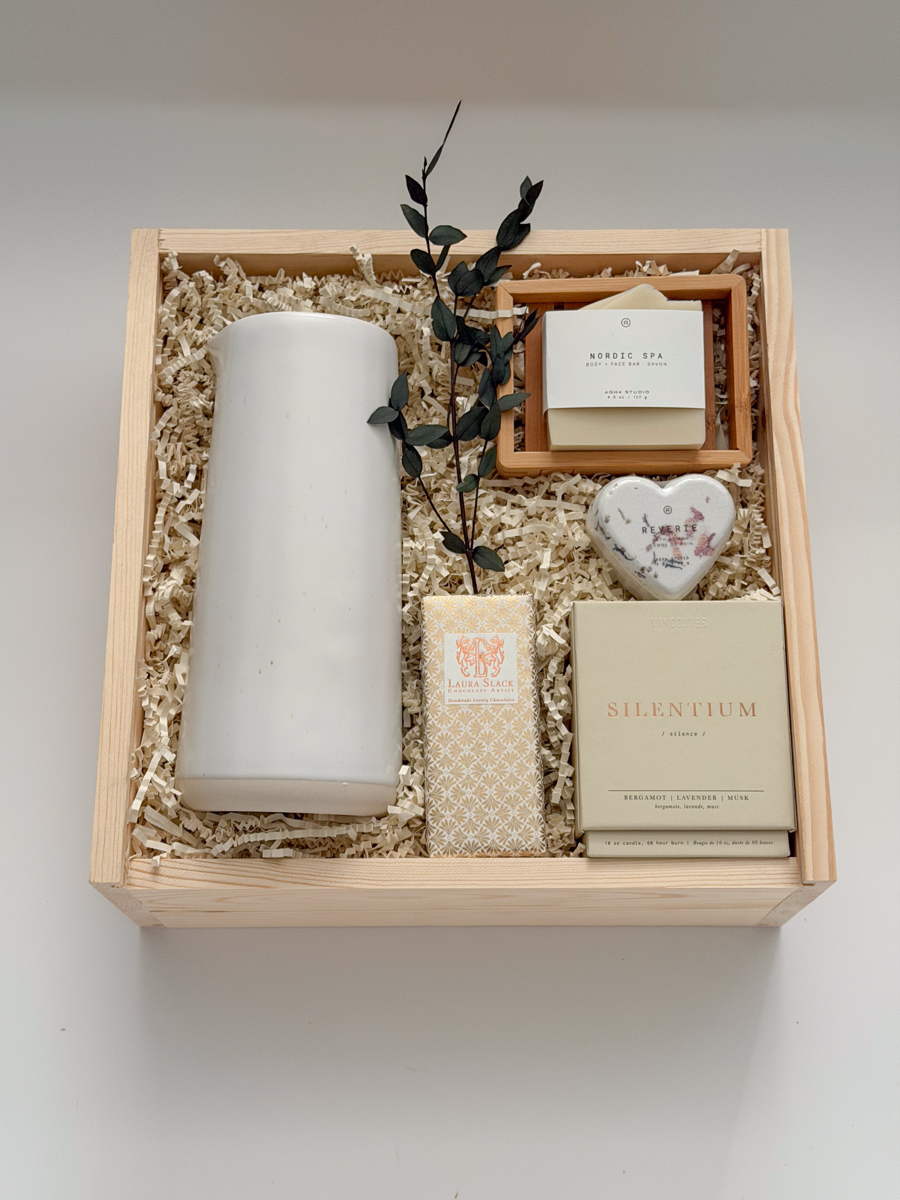 Overhead flat lay of The West Village by Muse Baskets and Co., showing a speckled white Fable carafe standing upright beside a Laura Slack artisan chocolate bar, a Vancouver Candle Co. Silentium candle, a Reverie heart bath bomb, and an Agha Studio Nordic Spa savon in a So Luxury bamboo dish, arranged in a Signature Luxe wood gift box. A luxury realtor closing gift and corporate gift.