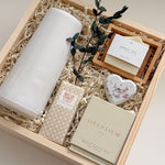 Close-up detail of The West Village gift box by Muse Baskets and Co., focused on a Laura Slack handmade chocolate bar in gold foil, a Vancouver Candle Co. Silentium candle in bergamot, lavender, and musk, a Reverie bath bomb, and an Agha Studio Nordic Spa savon, with the edge of a white Fable carafe and dried eucalyptus. A refined client appreciation gift for a new home.
