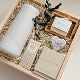 Close-up detail of The West Village gift box by Muse Baskets and Co., focused on a Laura Slack handmade chocolate bar in gold foil, a Vancouver Candle Co. Silentium candle in bergamot, lavender, and musk, a Reverie bath bomb, and an Agha Studio Nordic Spa savon, with the edge of a white Fable carafe and dried eucalyptus. A refined client appreciation gift for a new home.