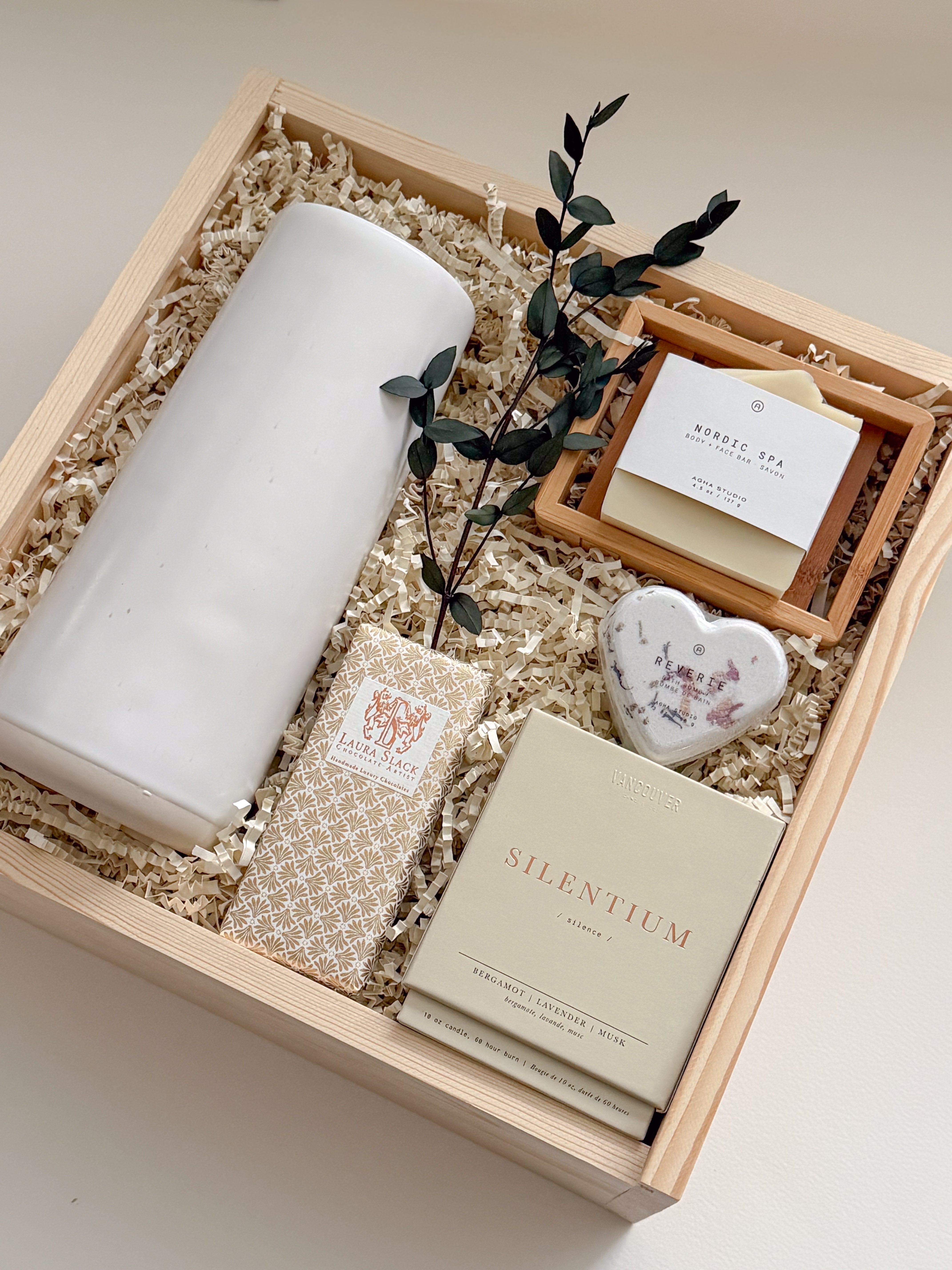 Close-up detail of The West Village gift box by Muse Baskets and Co., focused on a Laura Slack handmade chocolate bar in gold foil, a Vancouver Candle Co. Silentium candle in bergamot, lavender, and musk, a Reverie bath bomb, and an Agha Studio Nordic Spa savon, with the edge of a white Fable carafe and dried eucalyptus. A refined client appreciation gift for a new home.