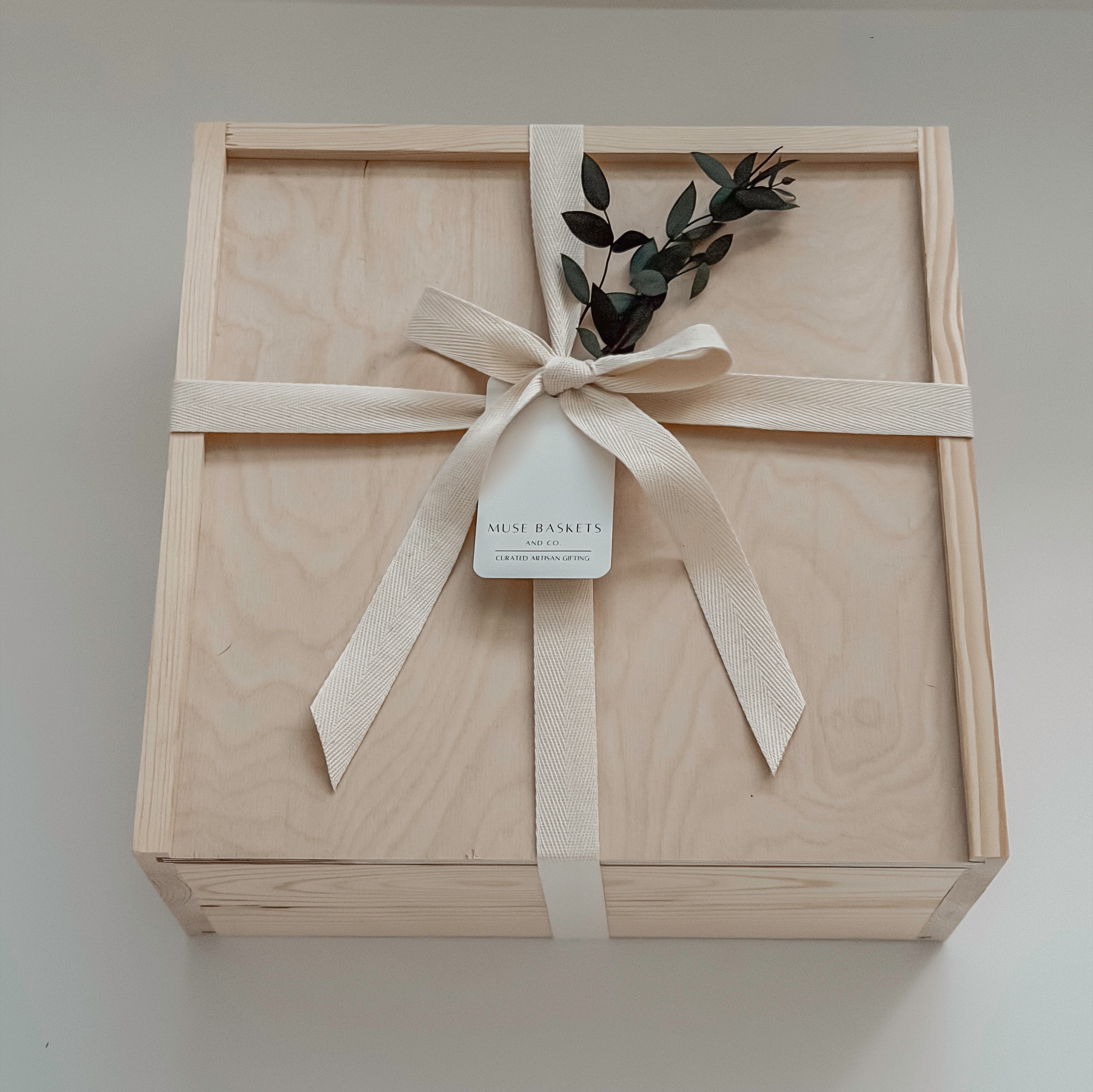 The West Village gift box from Muse Baskets and Co. shown closed, a Signature Luxe wood box tied with a cream cotton twill ribbon and dried eucalyptus, finished with a Muse Baskets and Co. gift tag reading Curated Artisan Gifting. A polished realtor closing gift, possession day gift, and corporate gift available for Vancouver, Lower Mainland, and Canada-wide delivery.