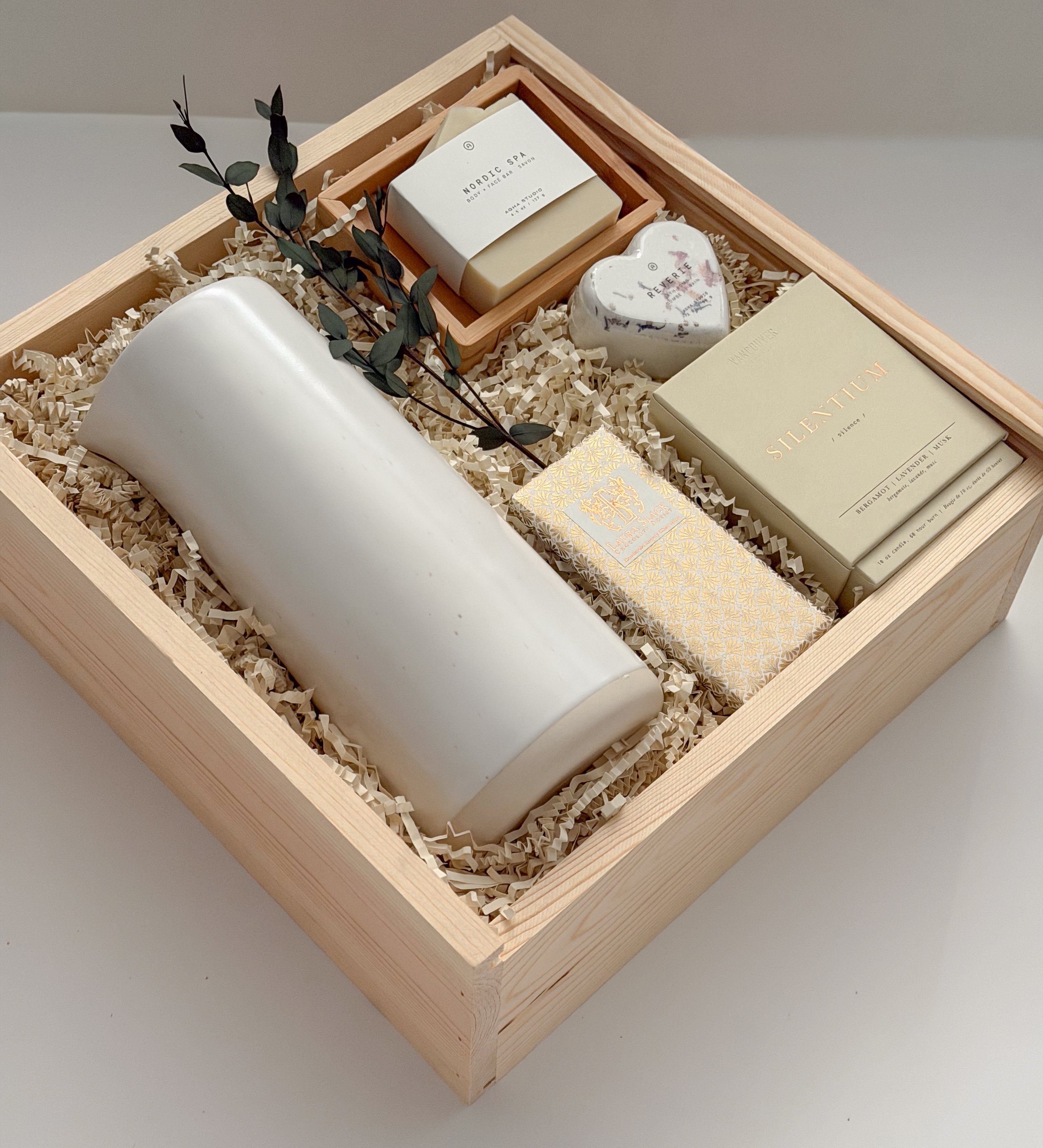 The West Village luxury gift box by Muse Baskets and Co. shown at an angle, holding a speckled white Fable carafe on its side, a Vancouver Candle Co. Silentium candle, a Laura Slack chocolate bar in gold Chiyogami wrapping, an Agha Studio Nordic Spa savon in a So Luxury bamboo dish, and a Reverie heart bath bomb with dried eucalyptus in a Signature Luxe wood box. A realtor closing gift and possession day gift for a new home.