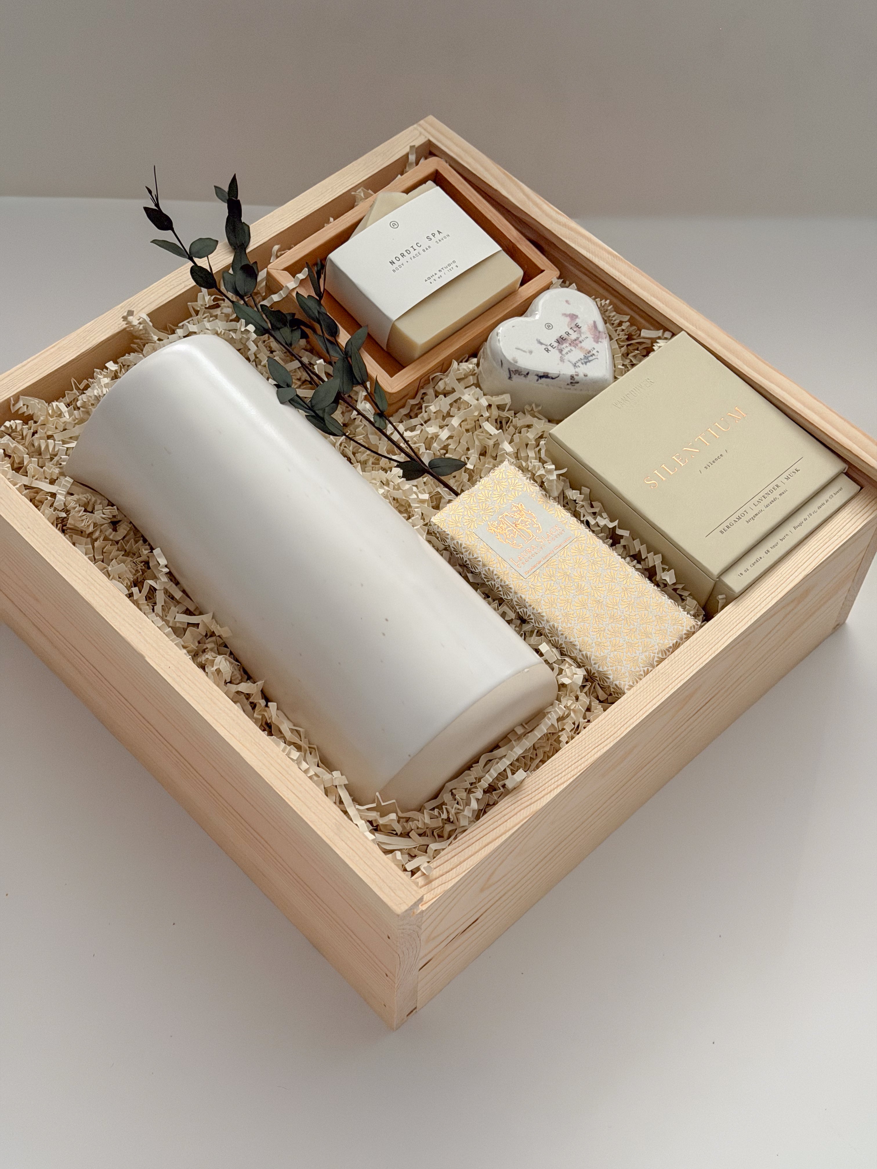 The West Village luxury gift box by Muse Baskets and Co. shown at an angle, holding a speckled white Fable carafe on its side, a Vancouver Candle Co. Silentium candle, a Laura Slack chocolate bar in gold Chiyogami wrapping, an Agha Studio Nordic Spa savon in a So Luxury bamboo dish, and a Reverie heart bath bomb with dried eucalyptus in a Signature Luxe wood box. A realtor closing gift and possession day gift for a new home.